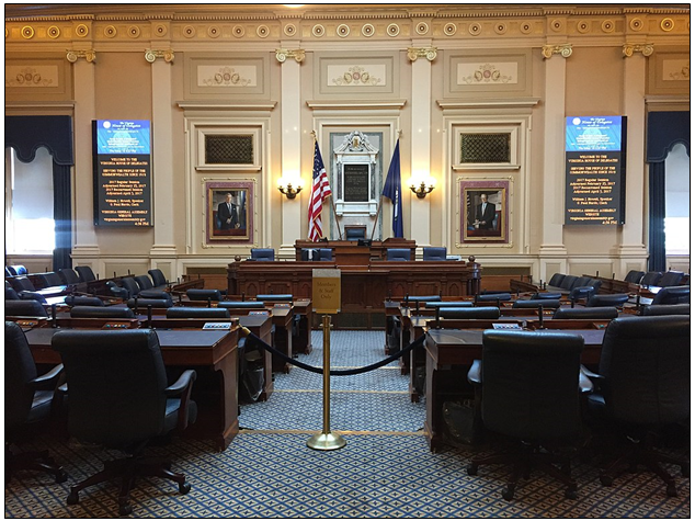 inside of meeting room in Virginia State Capital building. Several rows of desks and chairs point toward a central desk with the American and Virginian flag draped behind. On either side of the central desk two digital screens show upcoming information